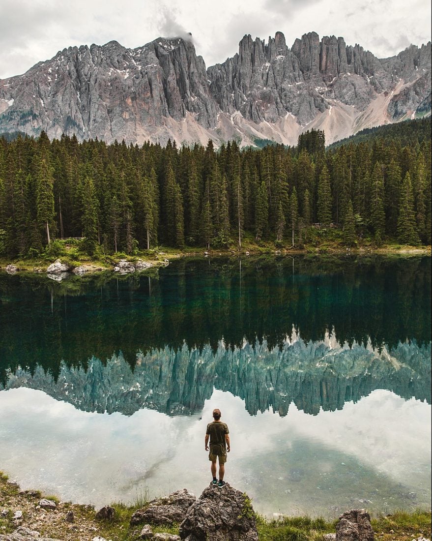 Man standing on a rock in front of a lake with mountains reflecting on it in the Dolomites, Italy.