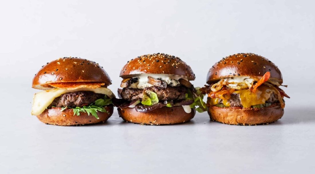 three cheeseburgers with different toppings arranged in a row against a white background